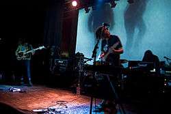 A band performing live on-stage. From left to right, a bassist, guitarist and drummer perform against a blue backdrop.