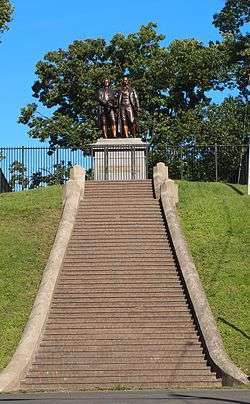 Photograph showing a long, broad flight of steps leading to a large metal statue of two men. There are about 50 steps. The two men in the statue are roughly life-sized, and are standing on a large stone pedestal. The pedestal has bronze plaque that reads "Goethe und Schiller".