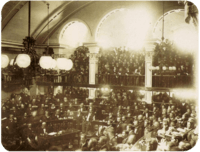 A photograph showing a large number of men seated on semi-circular tiers in a vaulted chamber as a large crowd looks on from an arcaded balcony