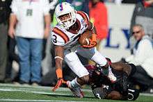 A receiver dressed in white with maroon and orange stripes is tackled by an opposing player in black and red.