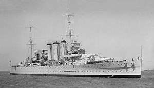 Photograph of a war ship in a body of water. Large cannons can be seen on the deck, along with sailors.