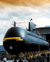 A submarine emerging from a large building. The submarine is decorated with flags, and the name "COLLINS" is painted across the front. There are a number of people around the base of the submarine.