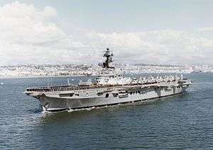 Colour photo of an aircraft carrier underway. Black-uniformed personnel line the perimeter of the flight deck, and a large number of aircraft with wings folded over sit on the deck.