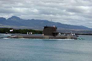 Side view of submarine with sailors on deck, at sea in front of a hilly coast