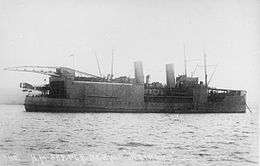 Black and white photograph of a ship. The vessel has two funnels, a large boxy superstructure topped by a crane, and an aircraft on her stern