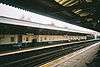 A railway platform with a railway track running from the bottom left corner to the middle right and the legs of several sitting people visible in the bottom right