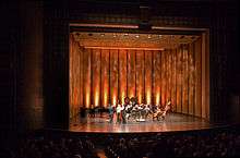 The lighted stage of a theatrical production, seen from the left side of the audience in a dark theater