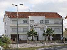 Headquarters of the Portuguese Badminton Federation. Three-story white building with balconies. Logo and "Federação Portuguesa de Badminton" written on tiles on façade.