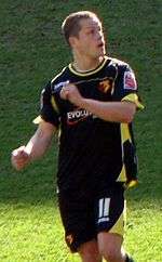 A man wearing black shirt and shorts with yellow trim, standing on a grass field.