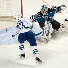 An ice hockey player wearing a white and blue jersey scores against a goaltender wearing a teal and black jersey with white pads.  The player has both hands on his stick outstretched to direct the puck in the net, while the goaltender is off balance looking back at the puck in his net.