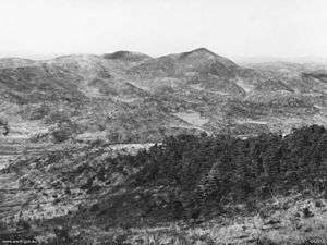 A series of ridgelines and steep hills in the distance, with Maryang San on the right. In the foreground is a heavily vegetated knoll, with a valley in the intervening ground.