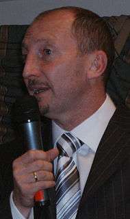 A man wearing a shirt, tie and jacket; holding a microphone.