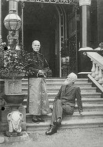  outdoor shot with two men on steps of a large house: bearded Western men (seated, right) wearing suit; one oriental-looking man (left, standing) wearing Chinese garments