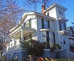 A white house with frilly rooflines lit by the sun from the left, seen from the left and slightly below. It has a tower on the right with a brick chimney.