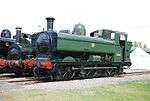 A green pannier tank locomotive, viewed from the left and front, is standing in front of two other locomotives. The locomotive is fitted with a chimney, rounded boiler dome, and a curved safety valve cover. The cab is enclosed with rectangular windows. Although mainly green, the chimney, front, and undercarriage are painted black, except the coupling rods which are grey, and the front buffer beam is painted red. On the side of the pannier tank is a yellow roundel with the letters G W R. On the side of the cab there is a small blue disc and a black number plate with the letters and edging in yellow.