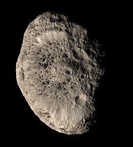 An irregularly shaped oblong body is illuminated from the left. The terminator is near the right limb. The body is elongated in the top-bottom direction. The surface is punctured by numerous impact craters, which make it look like a sponge or cheese.