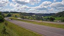 A divided expressway curving through a rural landscape alongside a valley, climbing slightly in the background.