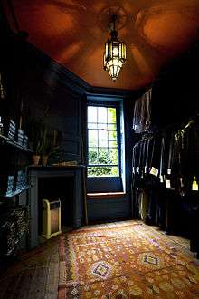 A wood panelled room, with polished wood floor, partly covered by a rug in the foreground. The room is lit by a gothic, stained glass ceiling light and from a single sash window. To the left of the window is a fireplace, with modern heater. The three shelves on the left wall are filled with shirtboxes. The right wall is hung with two rows of shirts and jackets, which have labels on their sleeves.