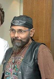 An bearded Indian man with spectacle standing, smiling, a black cap around his head