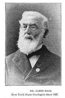 A black-and-white photographic portrait of a bearded, white-haired bespectacled man, shown from the chest up