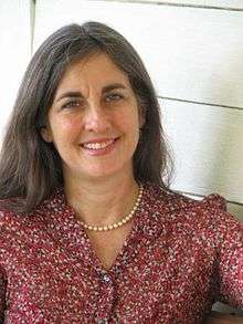 A smiling woman with slightly graying hair, in a maroon print blouse with a small string of pearls sits outside in front of a building.