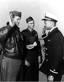 Three man stand on the deck of a ship, one with his arm raised and another reading from a paper.