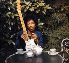 A color image of a man sitting at a table in the outdoors holding a black guitar.