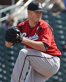 A man wearing a black baseball cap and a red jersey with "Sounds" written across the chest in white script and gray baseball pants shown preparing to pitch a ball from the mound