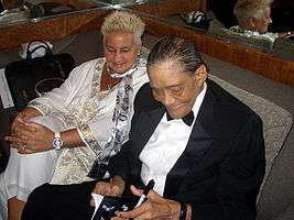 A man in a suit and a woman in a white outfit seated on a couch; both are looking toward reading material he is holding in his hands along with a pen.