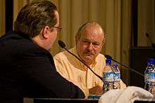 A middle-aged male in a white button-down shirt sits behind a microphone at a convention.