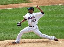 A man in a white baseball uniform with the word "Mets" written across the chest delivers a pitch.