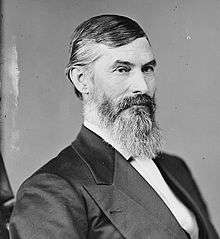 A man with graying hair, a mustache and a long beard wearing a dark jacket and tie and white shirt