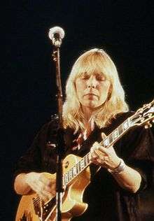 A blonde-haired woman stands in front of a microphone holding a guitar.