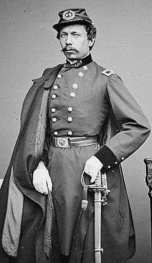 A man with curly hair and a Van Dyke mustache, wearing a forage cap, a long double-breasted jacket with belt, white gloves, and a coat hanging over his right shoulder. His left hand is holding a sword.