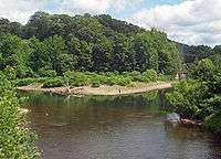 A body of water, seen from slightly above, that appears to fork in the center of the image. A metal bridge is partially visible crossing the right fork. The water in front is brownish, shading to green near the far shore. Some people are standing and fishing at the water's edge.