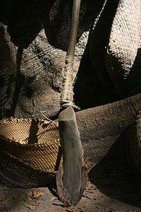 A metal blade attached to the base of a wooden rod resting against a tree trunk and various wicker products