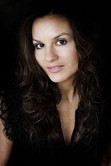 A light-skinned woman with long brown hair is seen looking in the direction of the camera.