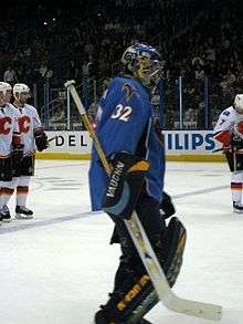 An ice hockey goaltender skates towards the right of the picture. He wears a blue jersey and blue helmet, with black leg pads and blocker