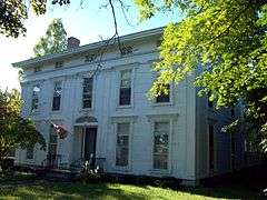 A white house seen from slightly to its right, with a tree branch partially obscuring it. Its front windows and door have some decorative features like columns. An American flag flies from a pole above the main entrance.