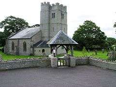 Stone building with square tower. In the foreground is a gateway with its own roof.