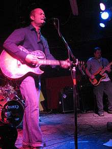 Young man singing and playing electric guitar on a stage with members of a rock band behind him