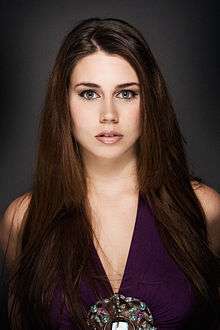 Photographic portrait of a young woman, looking directly to camera. She has long brown hair and is wearing a purple dress.