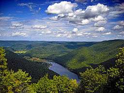 A view from atop a mountain of a blue lake between roling green hills and low mountains under a partly cloudy sky