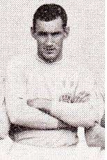 A man with short black hair, dressed in a light-coloured football shirt and light-coloured shorts, standing with his hands crossed.