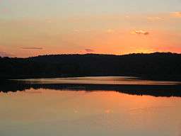 A red-orange sunset and the black silhouette of a line of trees are reflected in a smooth lake