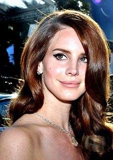 A fair-skinned young woman with long auburn hair and green eyes is looking to her right and smiling. She is wearing diamond ear studs, a diamond collier and her hair is in large curls.