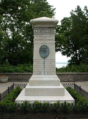 An upright stone monument surrounded by a low hedge.  In the middle is an upright oval of an elderly woamn's face in relief.