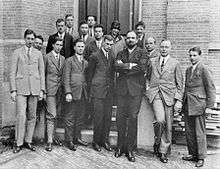 Fifteen men in suits, and one woman, pose for a group photograph