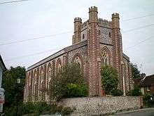 Large grey flint building with red brick trimmings, viewed from a slant so front and side are visible. There is a central tower with battlements at the front and from about two-thirds up, a roof slopes down each side. There are tall pointed-arch windows at the front and along the side. There is a grey wall in front of the building and the side is on the edge of an embankment.