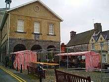 A couple of market stalls with red and white striped awnings in front of a stone building with three arched windows above stone arches